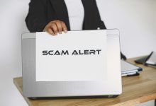 Pessoa segurando uma placa com o aviso “Scam Alert” diante de um laptop, representando um alerta contra fraudes online.