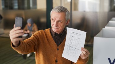 Eleitor idoso usa o celular para tirar uma selfie dentro de uma cabine de votação, segurando uma cédula.