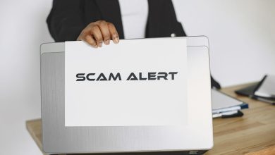 Pessoa segurando uma placa com o aviso “Scam Alert” diante de um laptop, representando um alerta contra fraudes online.