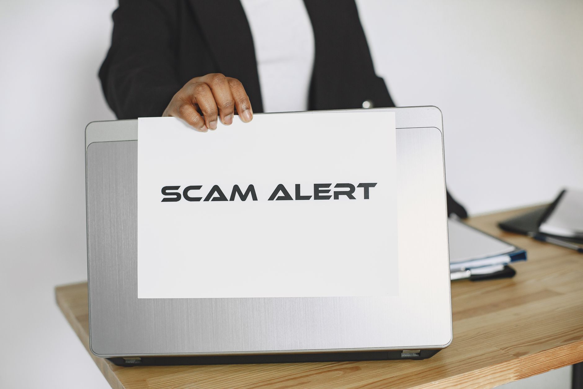 Pessoa segurando uma placa com o aviso “Scam Alert” diante de um laptop, representando um alerta contra fraudes online.