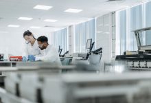 Dois pesquisadores trabalham em um laboratório de alta tecnologia usando óculos de proteção e luvas.