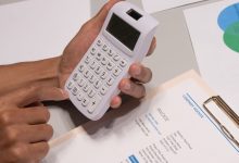 Mãos usando uma calculadora ao lado de uma fatura e documentos financeiros sobre uma mesa.