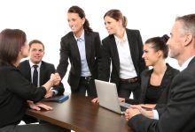 Grupo de pessoas reunido em um escritório, em uma conversa de trabalho, com participantes cumprimentando-se à mesa.
